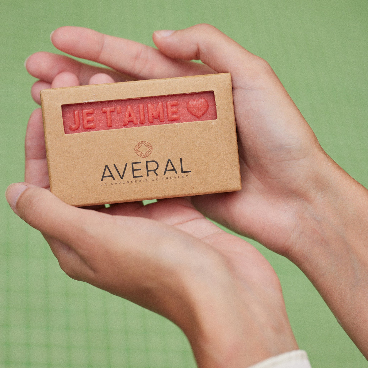 Hands holding a boxed Averal Provence Je t'aime French bar soap against a green background