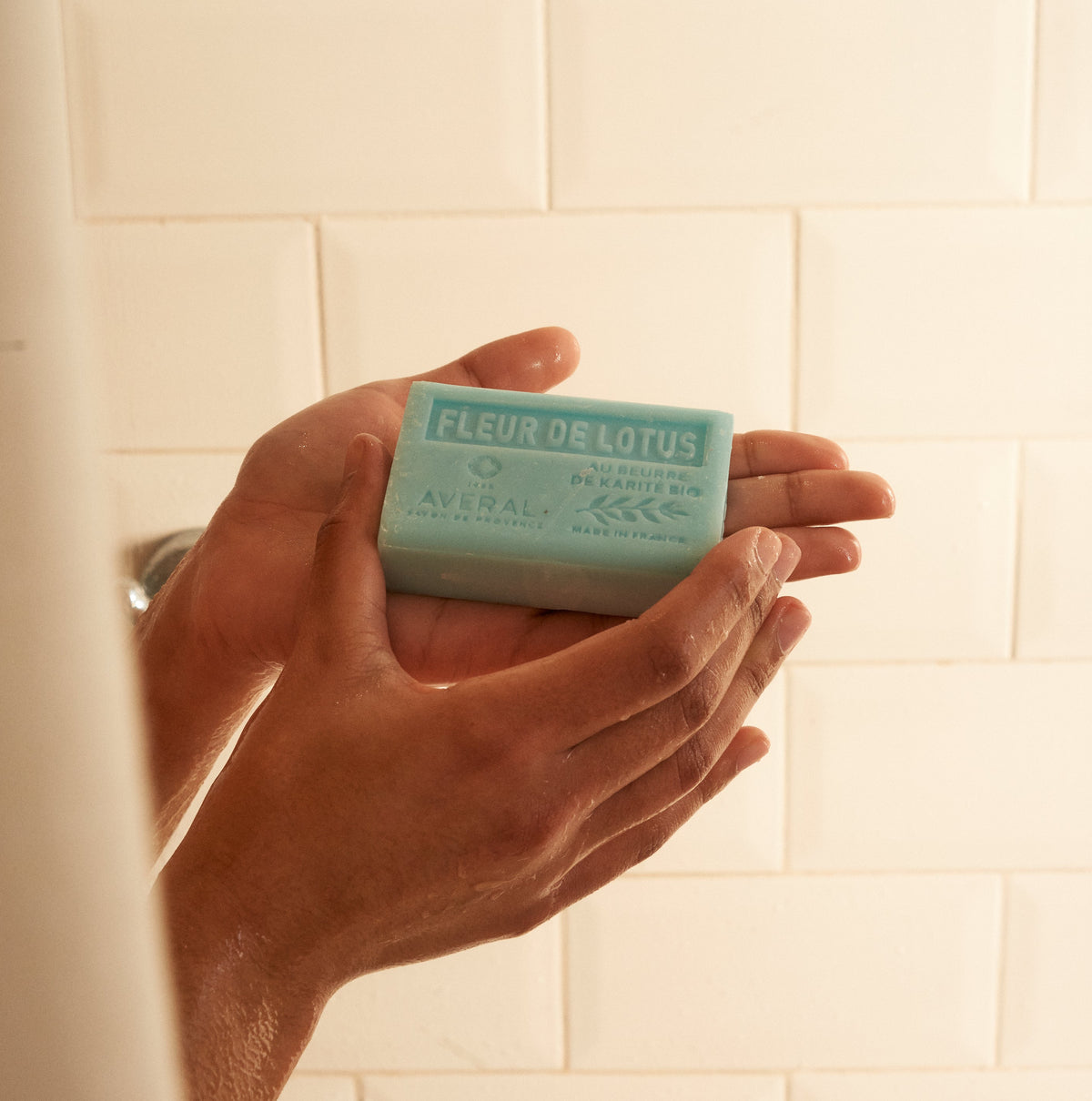 Open hand holding an Averal Provence Lotus Flower French bar soap in a cream tiled shower