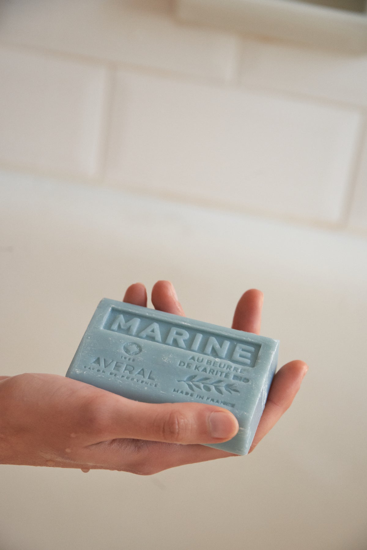 Hand holding a blue Averal Provence Marine French bar soap in front of white bathroom tiles