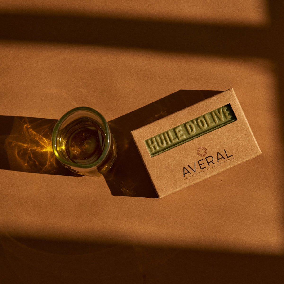 Averal Provence Olive Oil bar soap in its kraft box beside a glass of olive oil in warm light