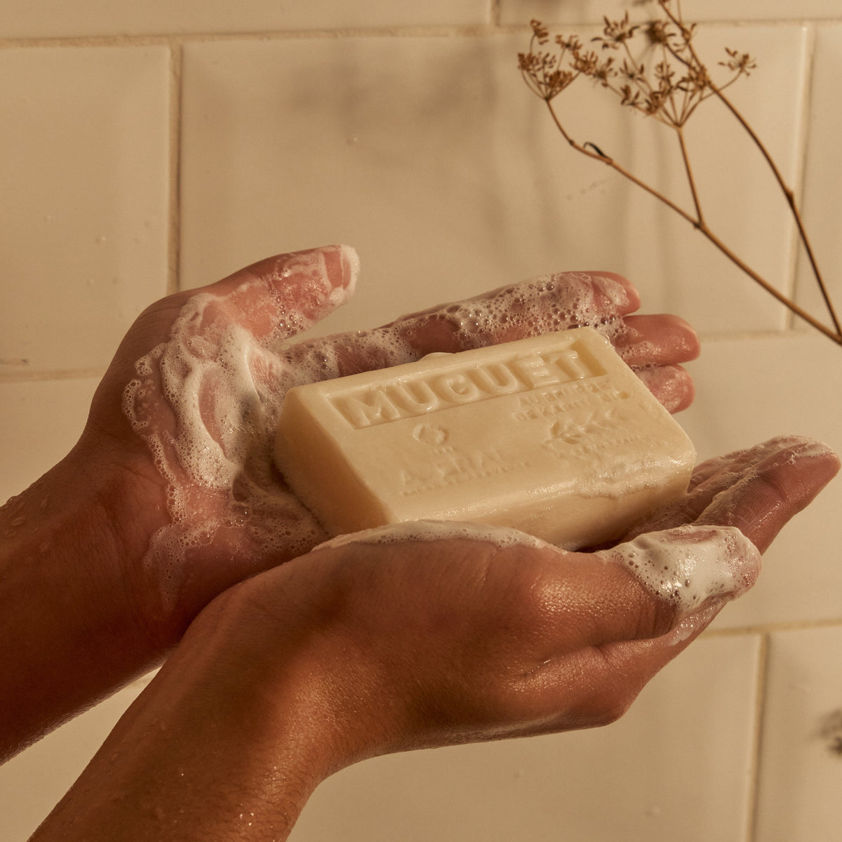 Lathered hands holding an Averal Provence Lily of the Valley French bar soap beside a dried flower stem